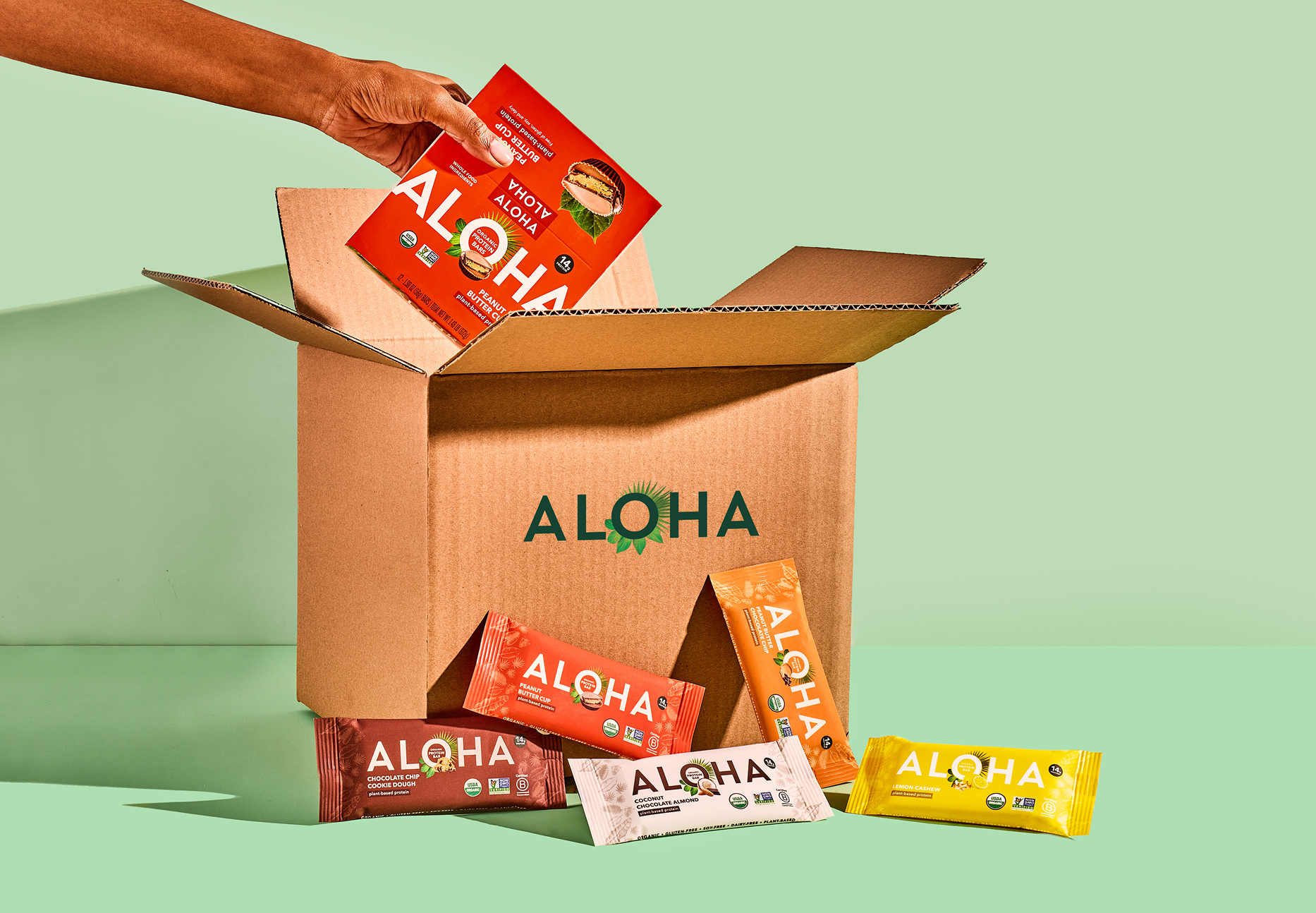 Hand placing Aloha protein bars into a cardboard box on a green background.